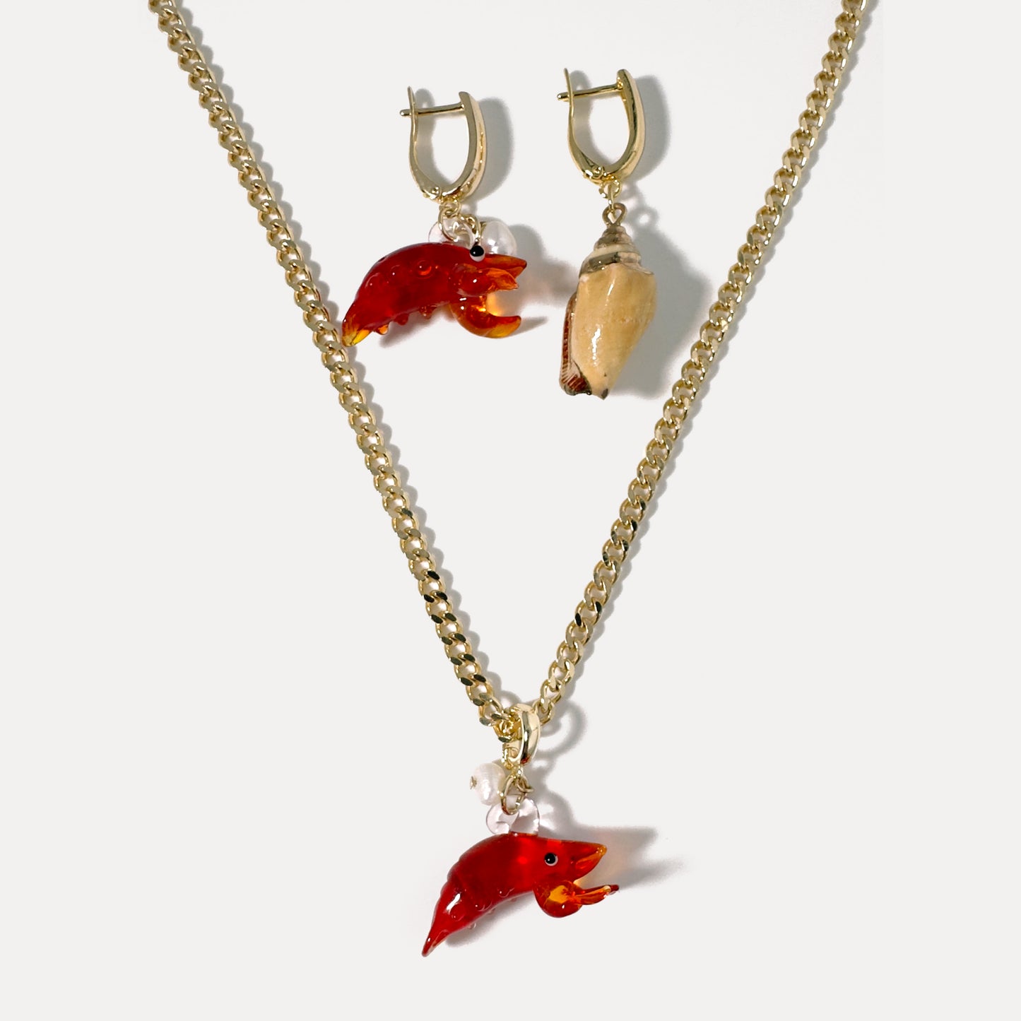 Glass lobster pendant necklace on 18K gold chain paired with asymmetrical 18K gold earrings — one featuring a glass lobster, the other a natural seashell